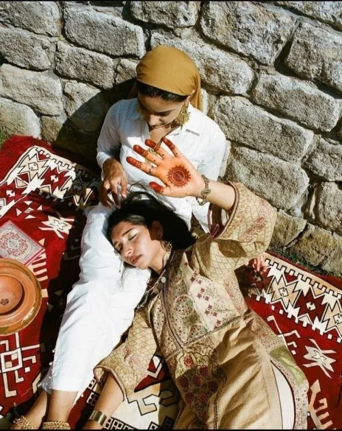 A woman with long black hair lying on a patterned rug against a stone wall, with her eyes closed and an intricate henna design on her palm. Another woman stands beside her, wearing a beige headscarf, white shirt, and glasses, looking down at her.