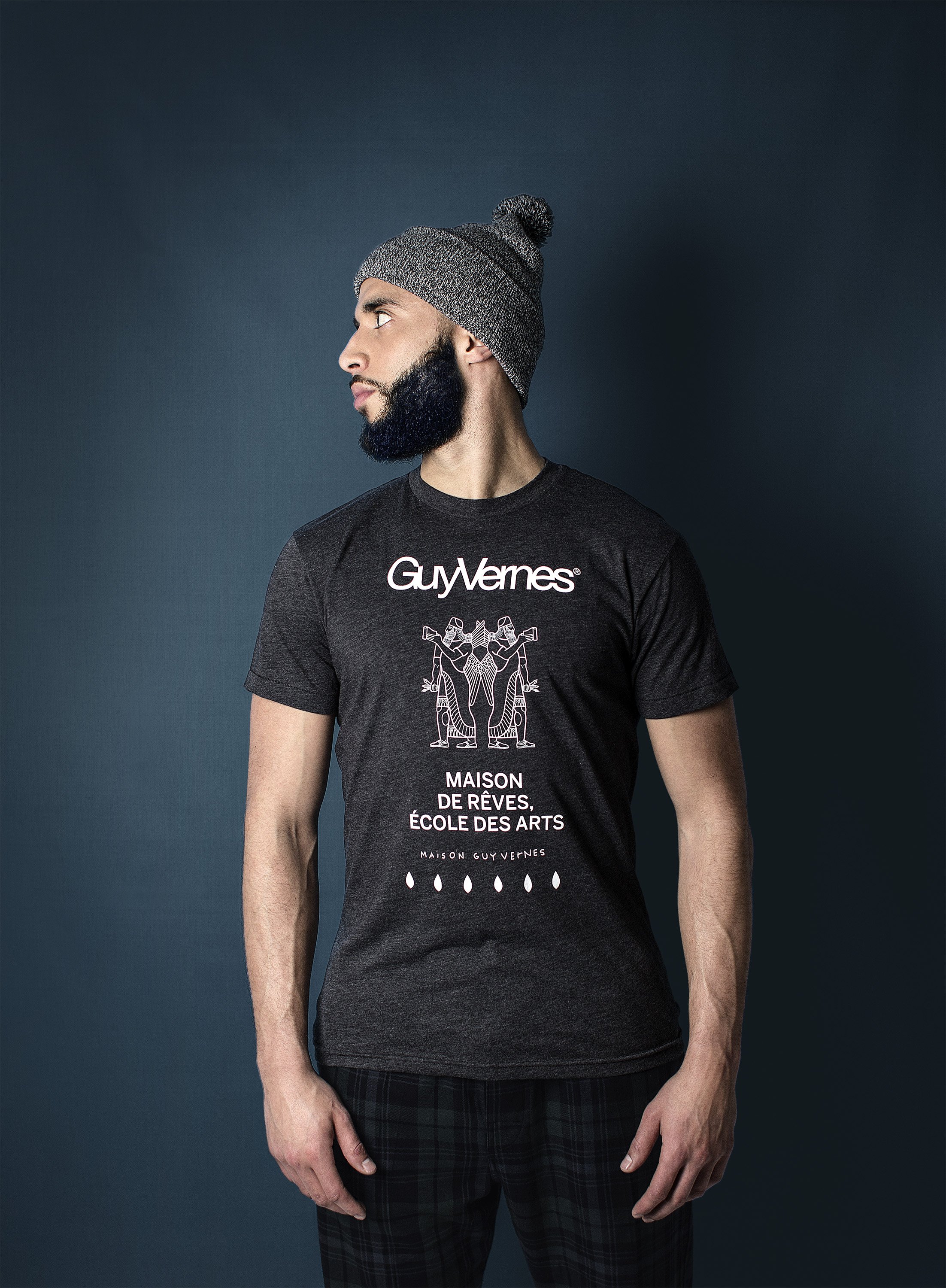 A man with a beard wearing a gray beanie hat and a black T-shirt with white text and graphics, standing against a dark blue background, looking to his left.