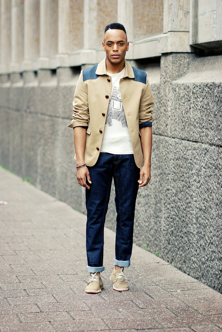 A young man standing on a sidewalk next to a stone building, wearing a beige jacket with dark shoulder patches, dark rolled-up jeans, and beige sneakers.