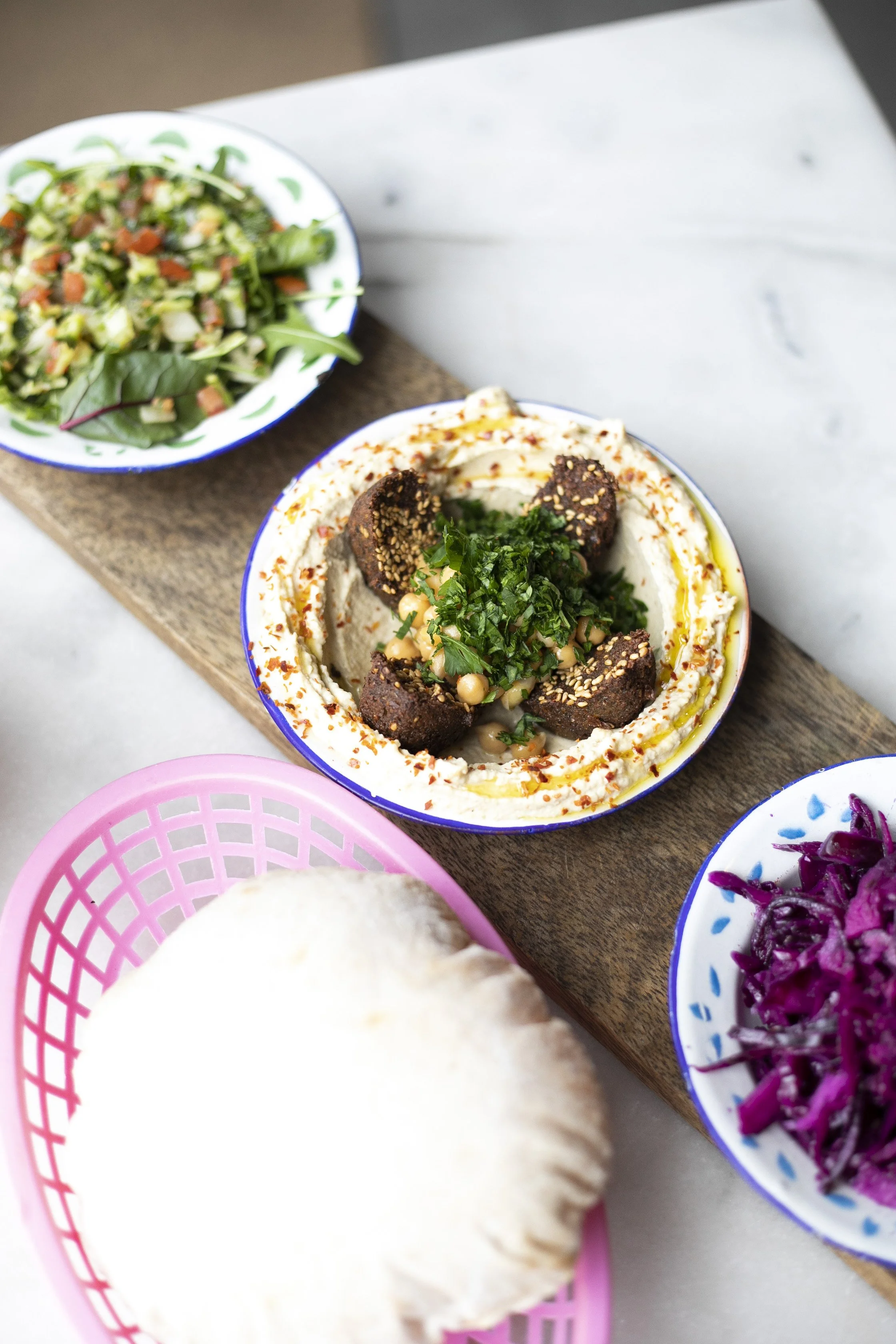 A wooden serving board featuring bowls of hummus topped with falafel, chopped herbs, and spices, a bowl of fresh salad with greens and vegetables, a basket of pita bread, and a bowl of purple shredded cabbage.