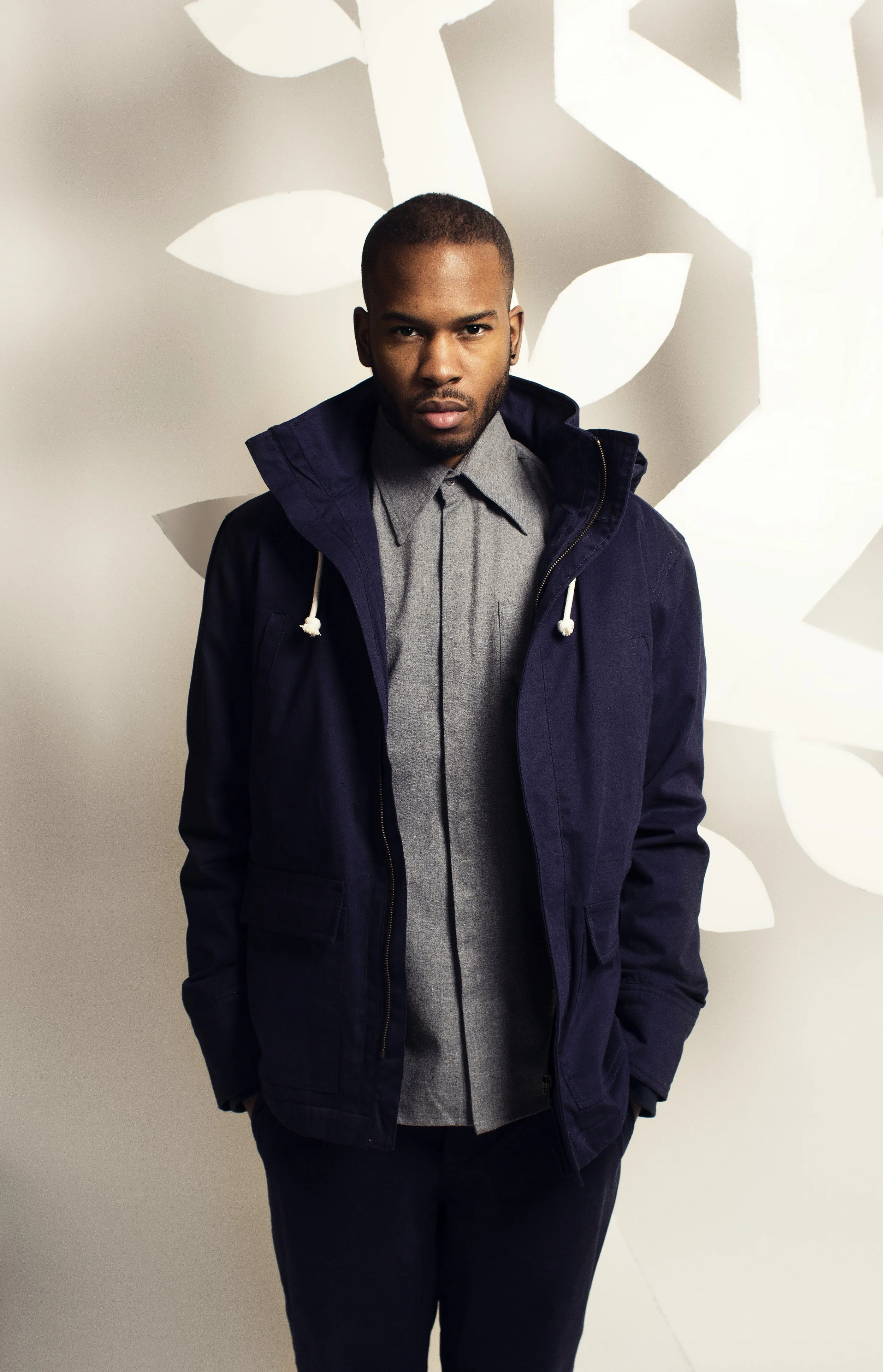 A young man standing in front of a white wall with large abstract leaf patterns. He is wearing a dark blue jacket over a gray button-up shirt and has a serious expression.