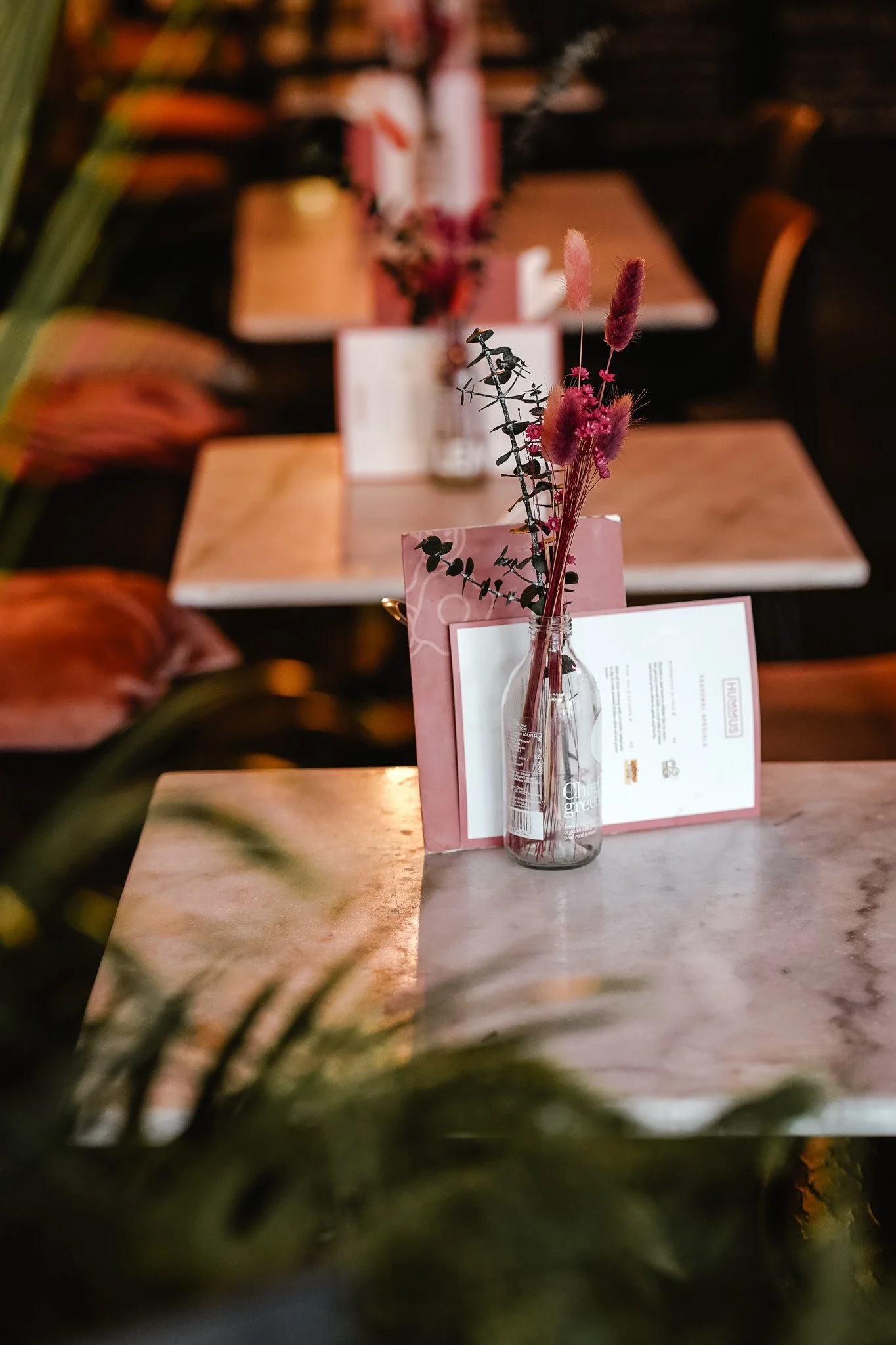 A table setting with a small glass bottle holding pink and purple dried flowers, placed on a pink and white menu or card in a row of similar tables.