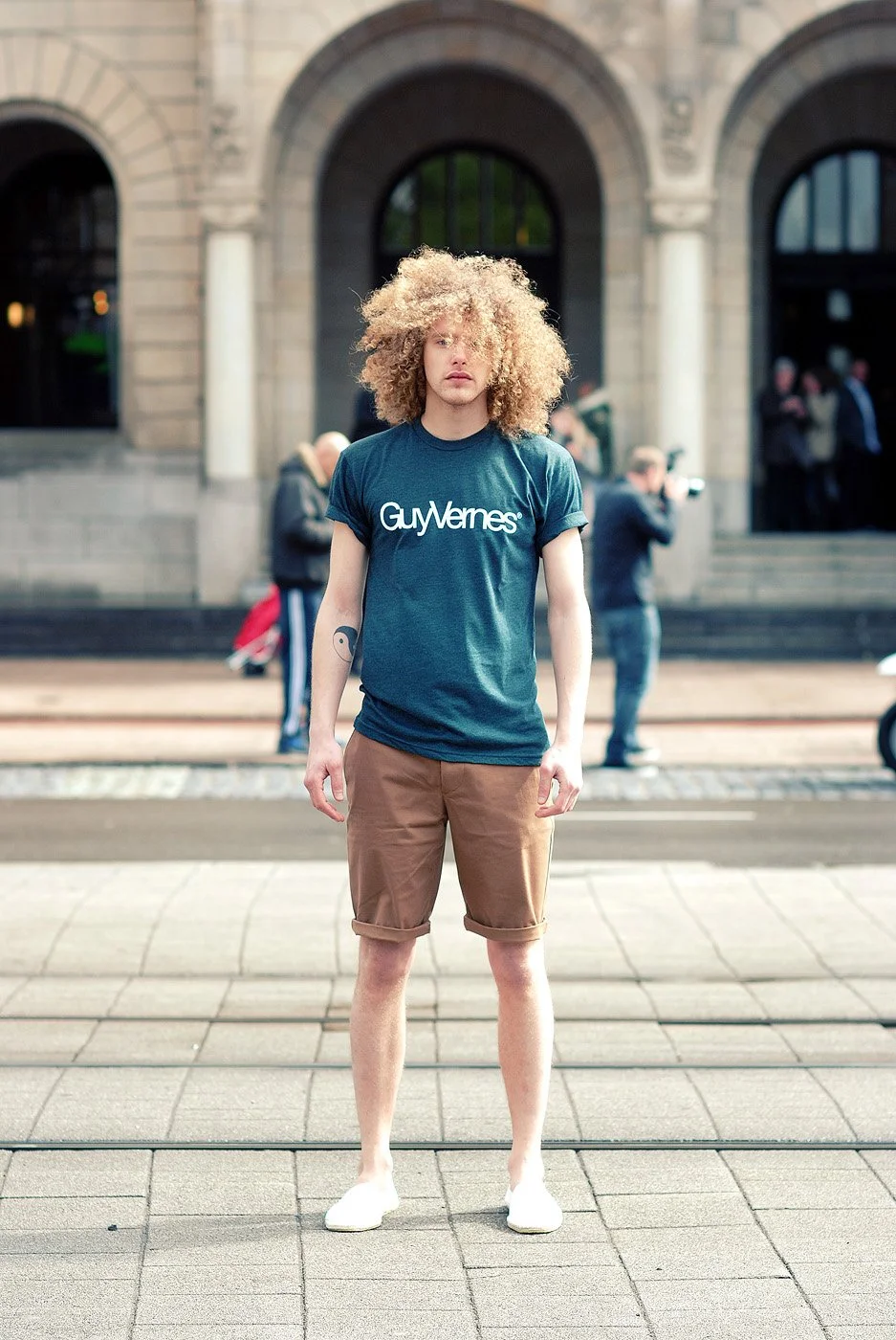 Young man with curly hair standing on a city sidewalk in front of a historic building, wearing a blue T-shirt and shorts.