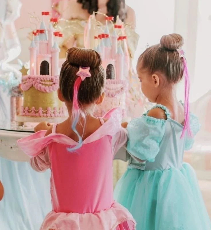 Two young girls dressed as princesses with colorful hair and princess costumes, looking at a castle-shaped birthday cake.