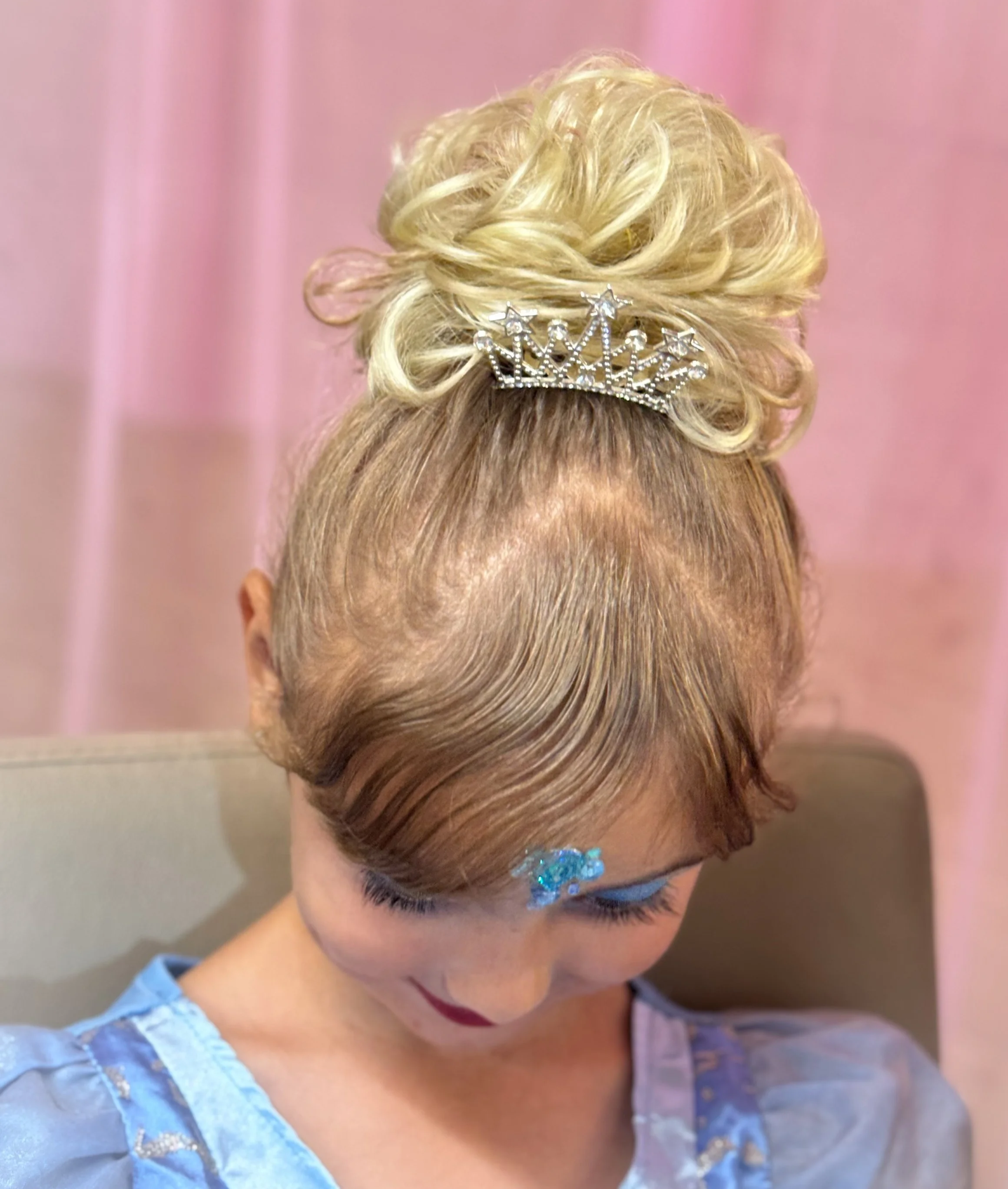A woman with elaborate blonde curled hair styled in an updo, wearing a small tiara, with blue, white, and silver makeup, traditional costume, against pink curtains.