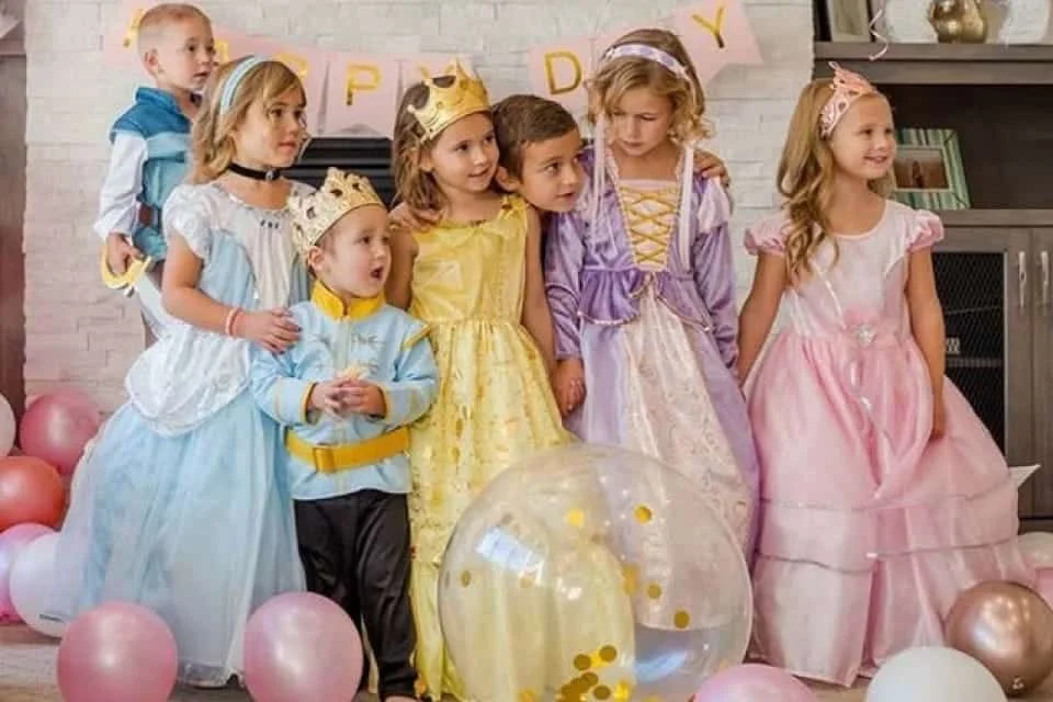 Six children dressed in princess, prince, and fairy costumes stand together at a birthday party with pink, white, and gold balloons and a decorated background that says 'Happy Birthday'.