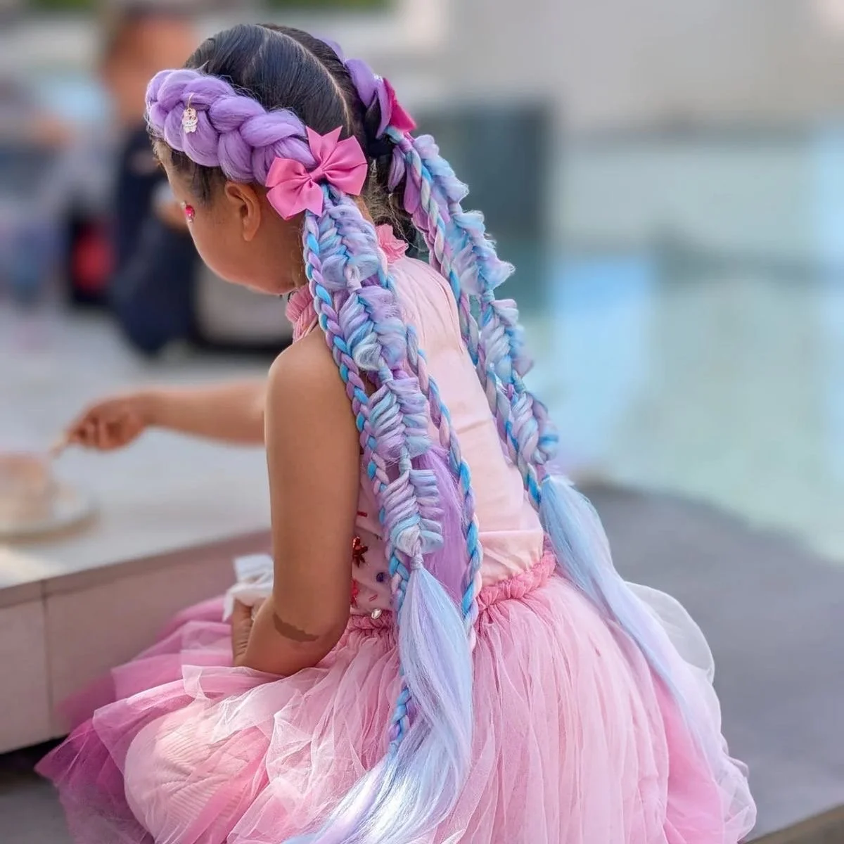 A young girl with long, braided hair in pastel purple, pink, and blue, decorated with pink bows, wearing a pink tulle dress.