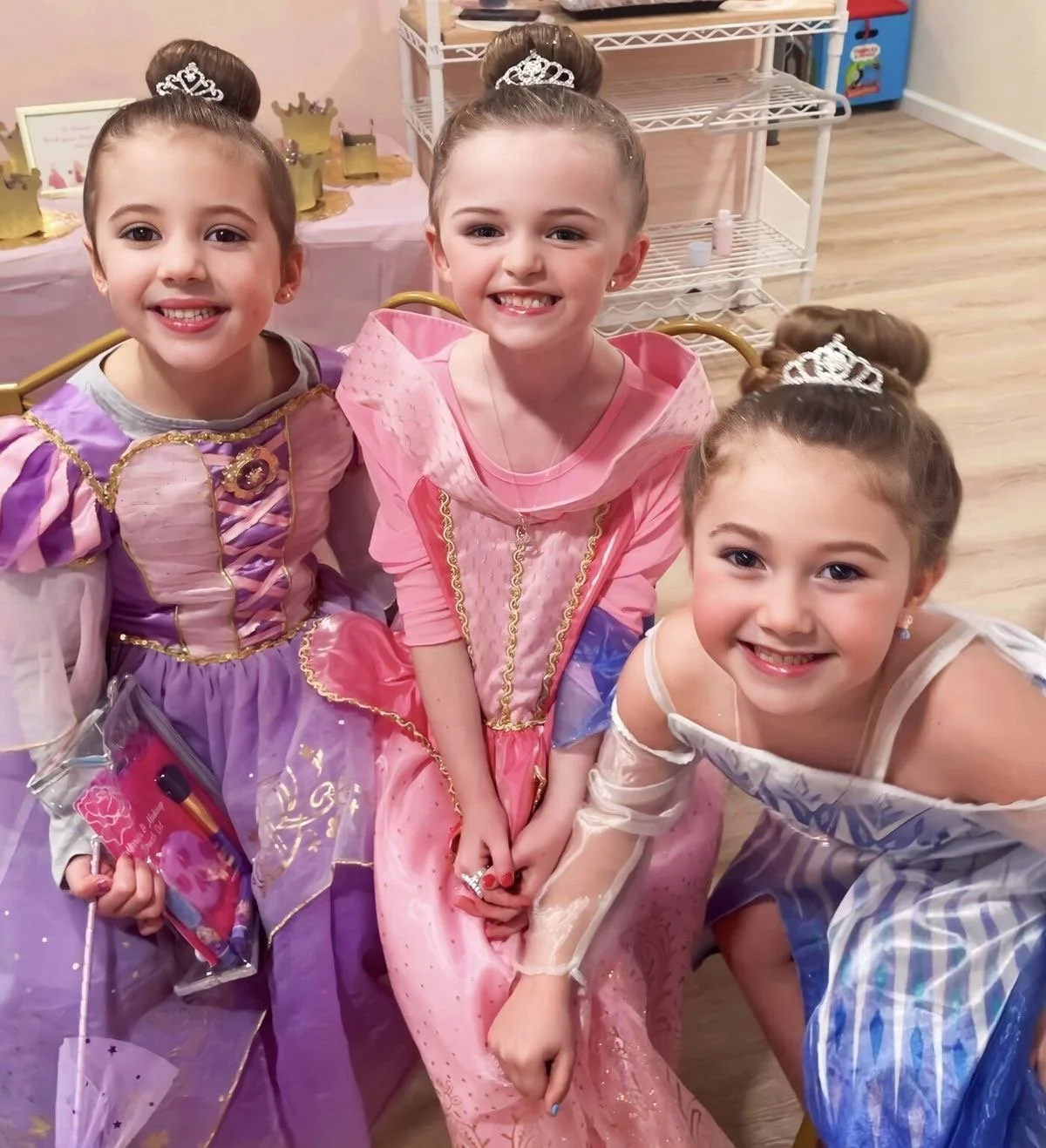 Three young girls dressed as princesses smiling at the camera in a decorated room with princess-themed decorations and a table in the background.