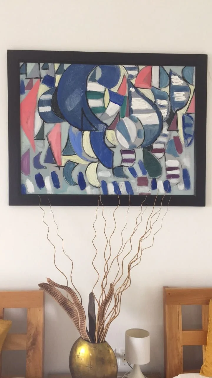 Abstract painting with various blue, red, green, purple, and white shapes and patterns hanging above a vase with decorative dried plant stems and leaves, and a small white lamp on a wooden surface.