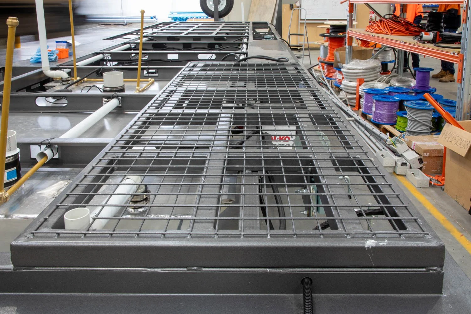 Industrial workspace with large stainless steel equipment and pipes, possibly part of a manufacturing or plumbing system, with shelves of cable spools and tools in the background.