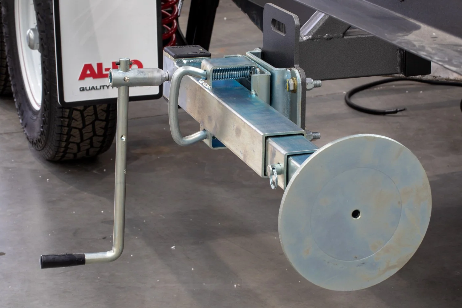 Close-up of a trailer hitch with a safety pin attached, connected to a metal support arm on a vehicle, with a tire and part of a white sign in the background.