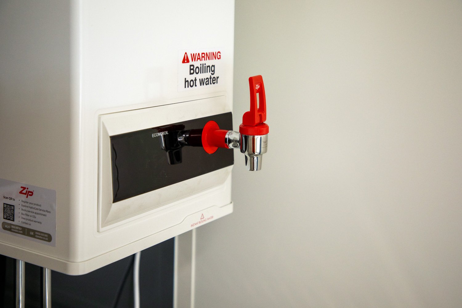 White water heater with a red and silver faucet, a warning sign that reads 'WARNING Boiling hot water', and a black panel on the front.