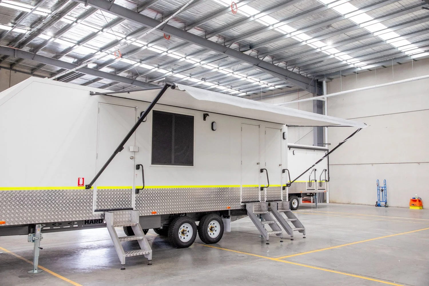 Inside a large warehouse with high ceilings. There are two mobile trailer units with stairs, black handrails, and retractable awnings. The floor is marked with yellow lines. A blue hand truck and an orange safety cone are in the background.