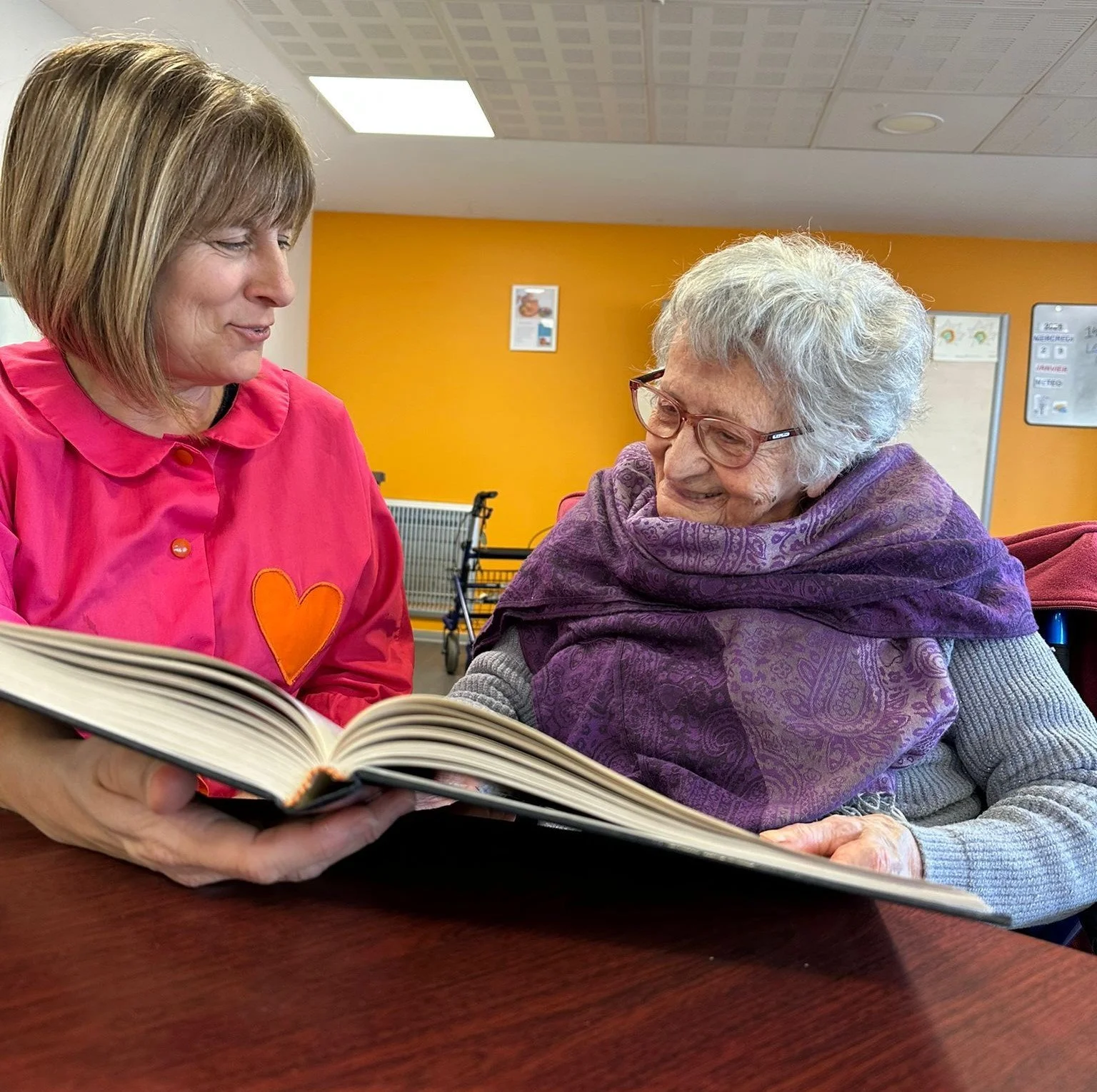 Une femme âgée avec des lunettes, assise à une table, regardant un livre avec une femme plus jeune qui lui montre le contenu, dans un environnement intérieur avec un mur orange en arrière-plan.