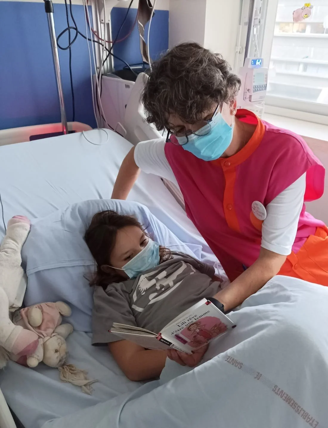 Une infirmière parle à une jeune fille hospitalisée dans un lit d'hôpital, toutes deux portent des masques faciaux, la jeune fille lit un livre.
