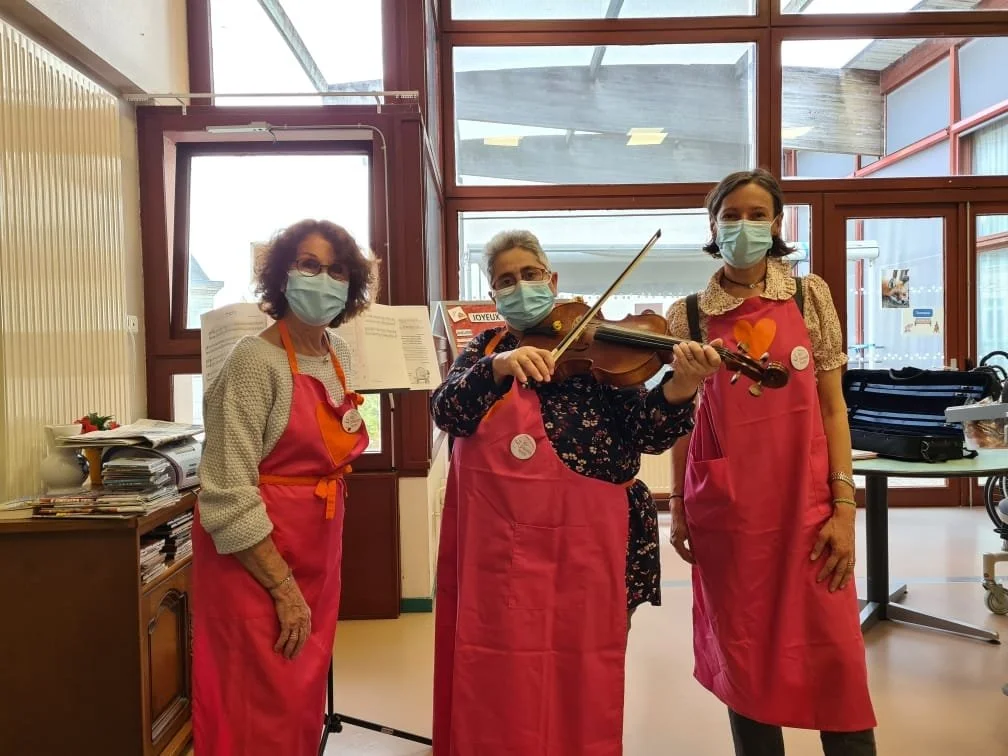 Trois femmes portant des masques buccaux, vêtues d'aprons roses, posent dans une pièce avec un violon, dans une atmosphère chaleureuse.