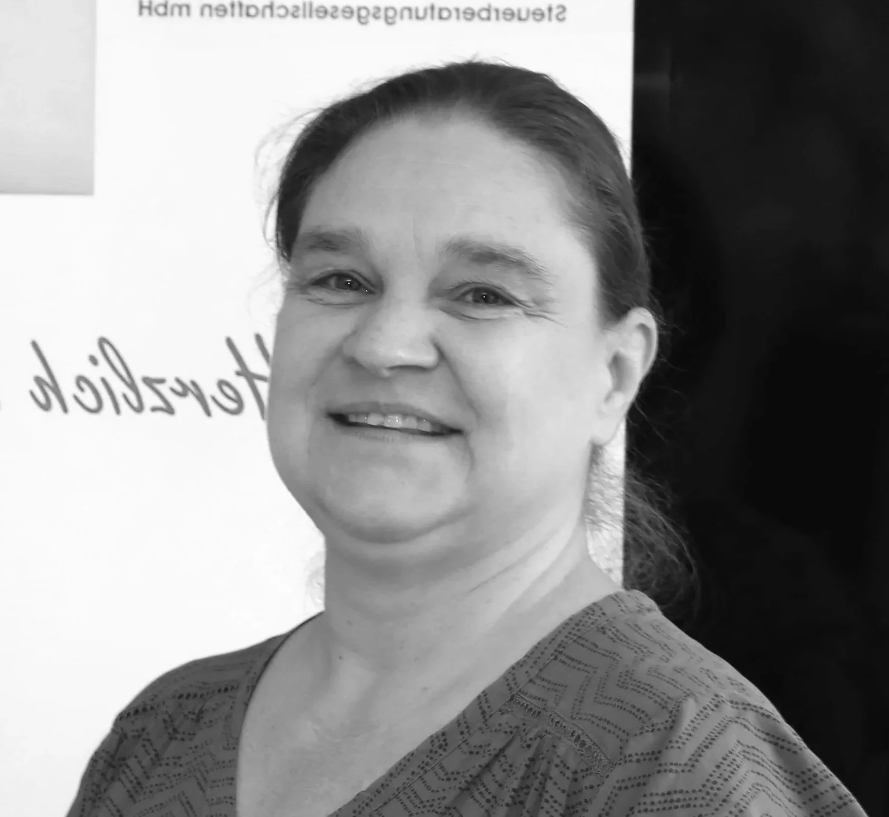 Schwarz-weiß-Fotografie einer lächelnden Frau mit dunklen Haaren, die eine Musterbluse trägt, vor einer weißen Wand mit Text.