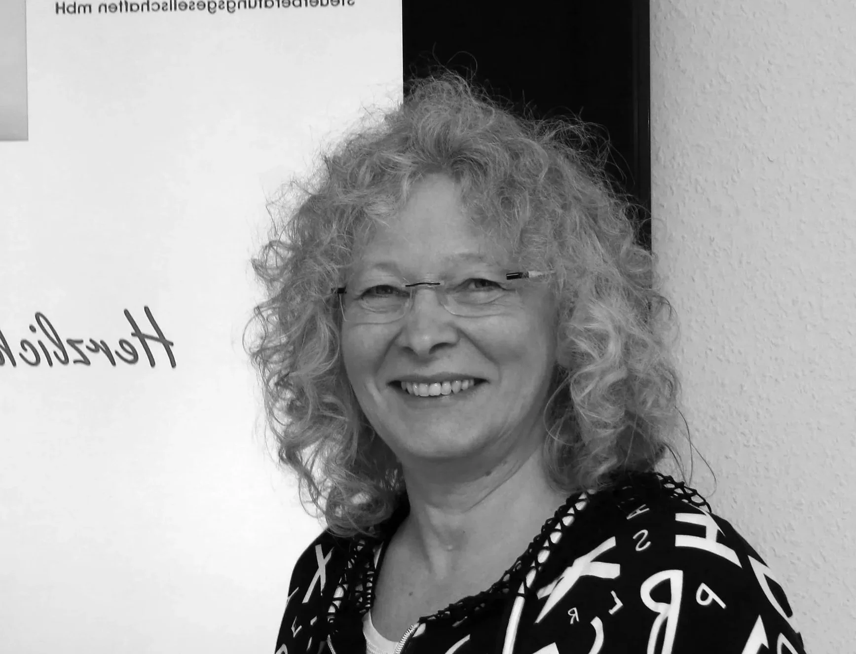 Schwarz-Weiß-Foto einer lächelnden Frau mit lockigem Haar und Brille, die vor einer Wand steht.