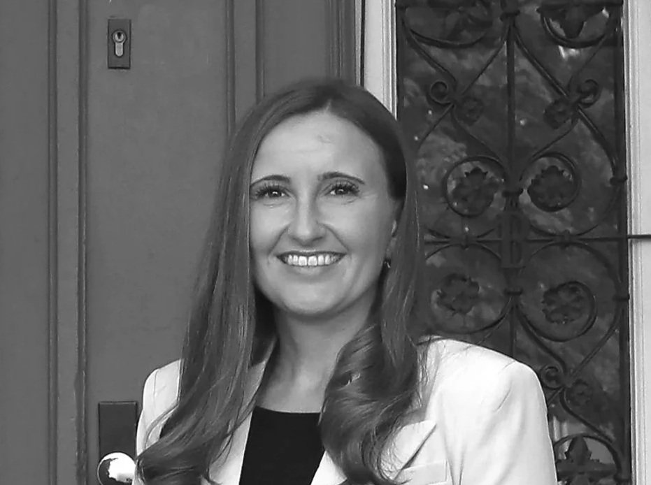 Ein lachender Mann mit langen Haaren in einem weißen Mantel steht vor einer Holz- und Metalltür.