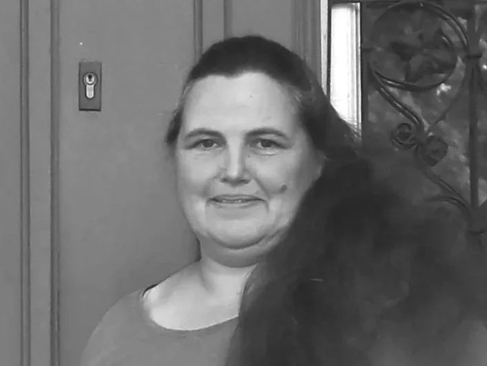 Schwarz-weiße Fotografie einer Person mit langen Haaren, die vor einer Wand mit dekorativem Gitter steht.