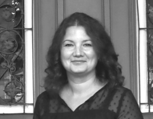 Schwarz-Weiß-Foto einer Frau vor einem Holz- und Glaseingang mit schmiedeeisernen Fenstern.