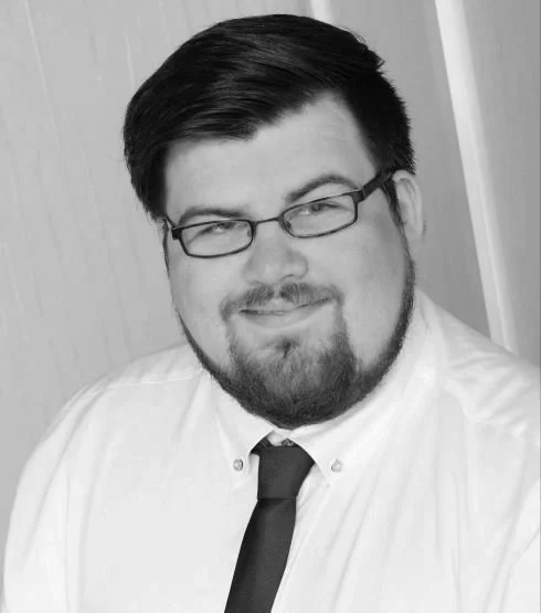 Schwarz-weiß Portrait eines Mannes mit dunklen Haaren, Bart, Brille, in weißem Hemd und schwarzer Krawatte, lächelnd.