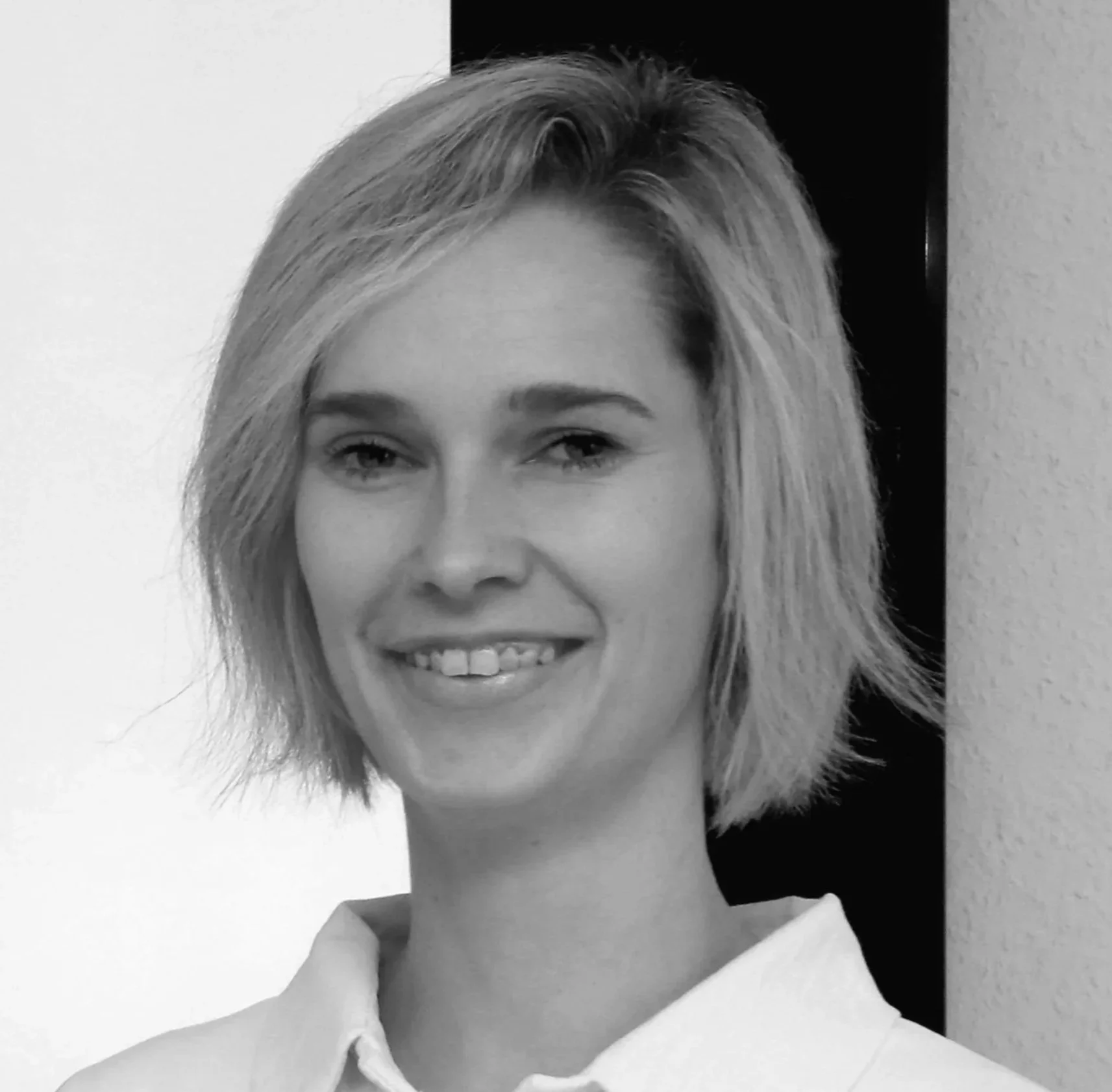 Schwarz-weiß Portrait einer lachenden jungen Frau mit kurzen, zerzaustem Haar, trägt eine weiße Bluse, vor einer Wand.