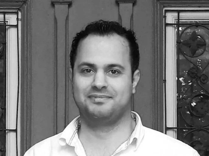 Ein Mann mit dunklen Haaren, weißem Hemd, vor einer Holzwand mit Fenster im Hintergrund.