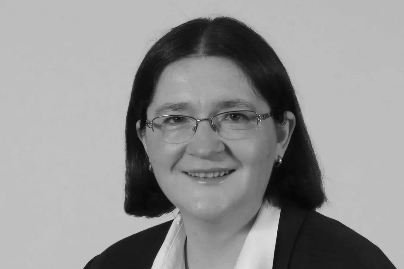 Schwarz-Weiß-Porträt einer Frau mit dunklen Haaren, Brille, schwarzem Blazer und weißen Hemd, lächelnd gegen einen hellen Hintergrund.