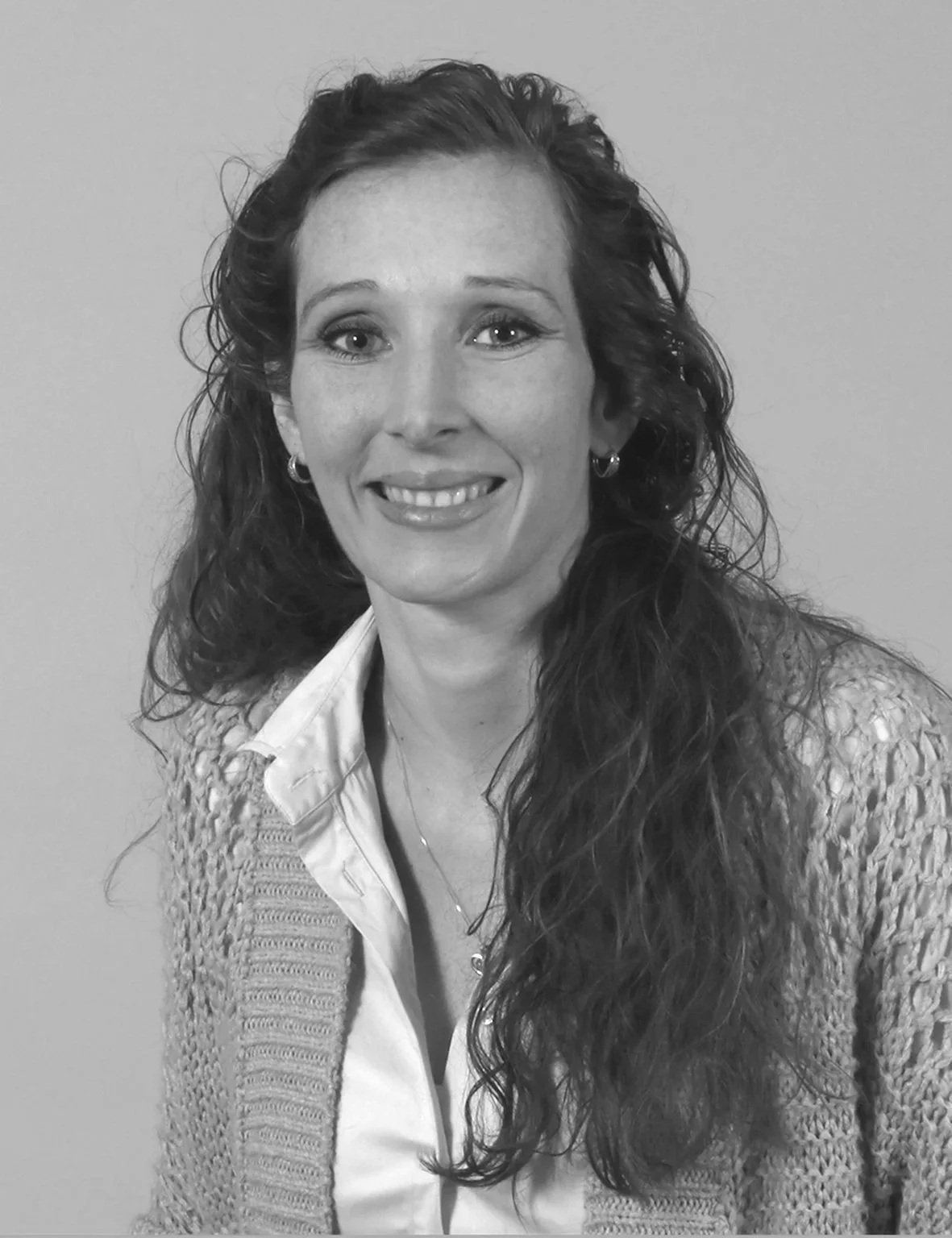 Schwarzweißfoto einer lächelnden Frau mit langen, lockigen Haaren, die eine weiße Bluse und eine gestrickte Jacke trägt.