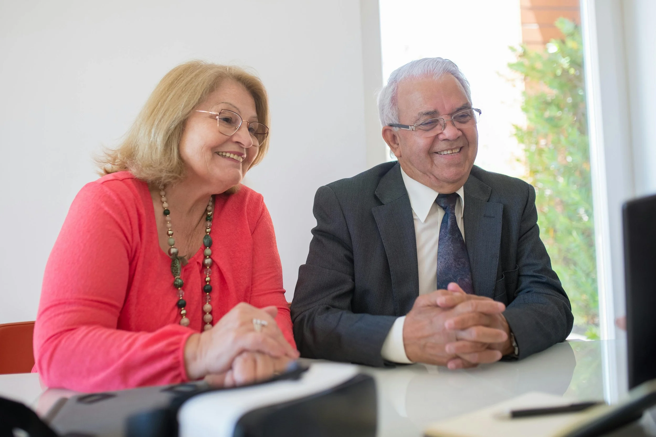 Zwei ältere Menschen, eine Frau und ein Mann, sitzen bei einem Meeting, lachen und wirken glücklich, mit einem Fenster im Hintergrund, durch das grüne Bäume sichtbar sind.