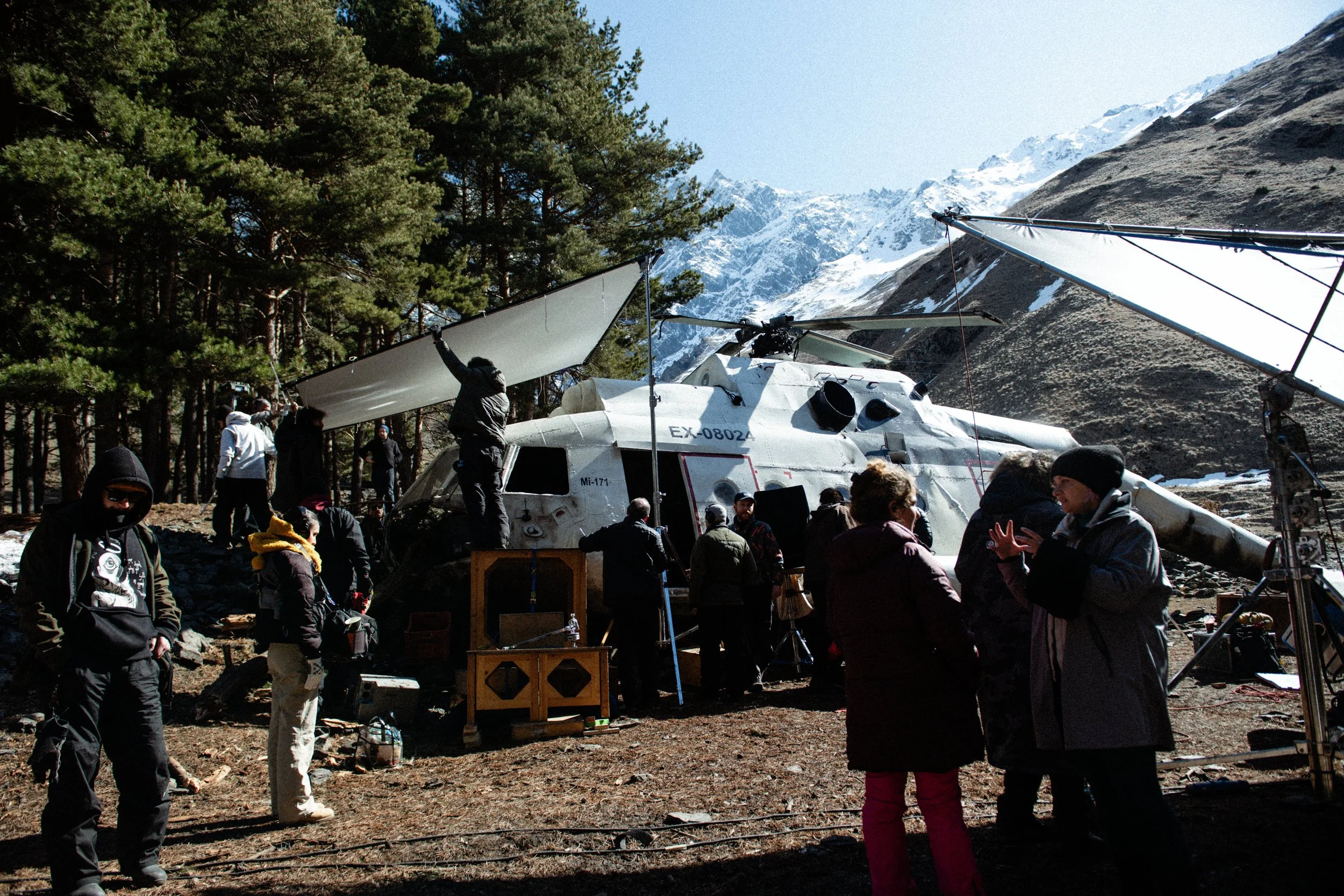 A film crew setting up a helicopter with a propeller blade attached, amidst snow-capped mountains and pine trees, while crew members and actors prepare for a shoot.