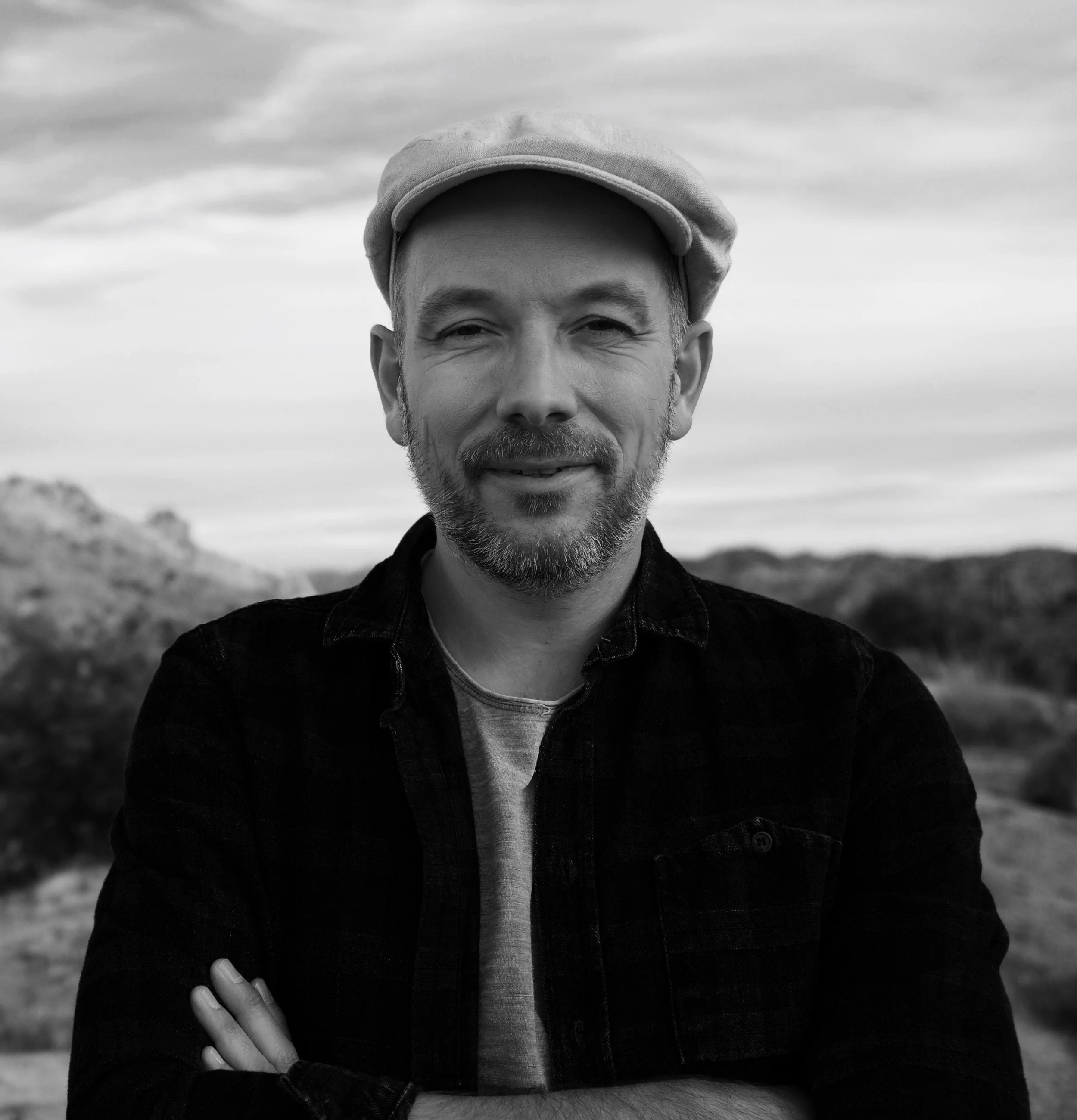 A man with a beard and mustache wearing a flat cap, a dark jacket, and a T-shirt, standing outdoors with mountains and sky in the background.
