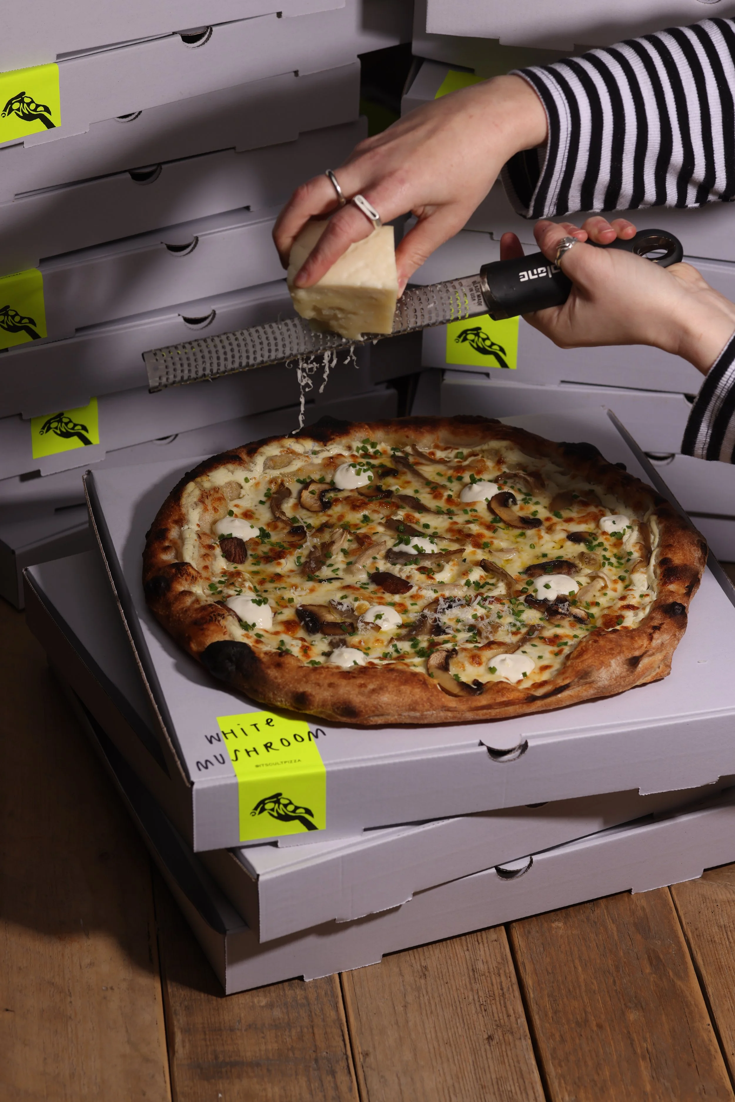 person grating fresh pecorino over a wild mushroom pizza on a stack of pizza boxes