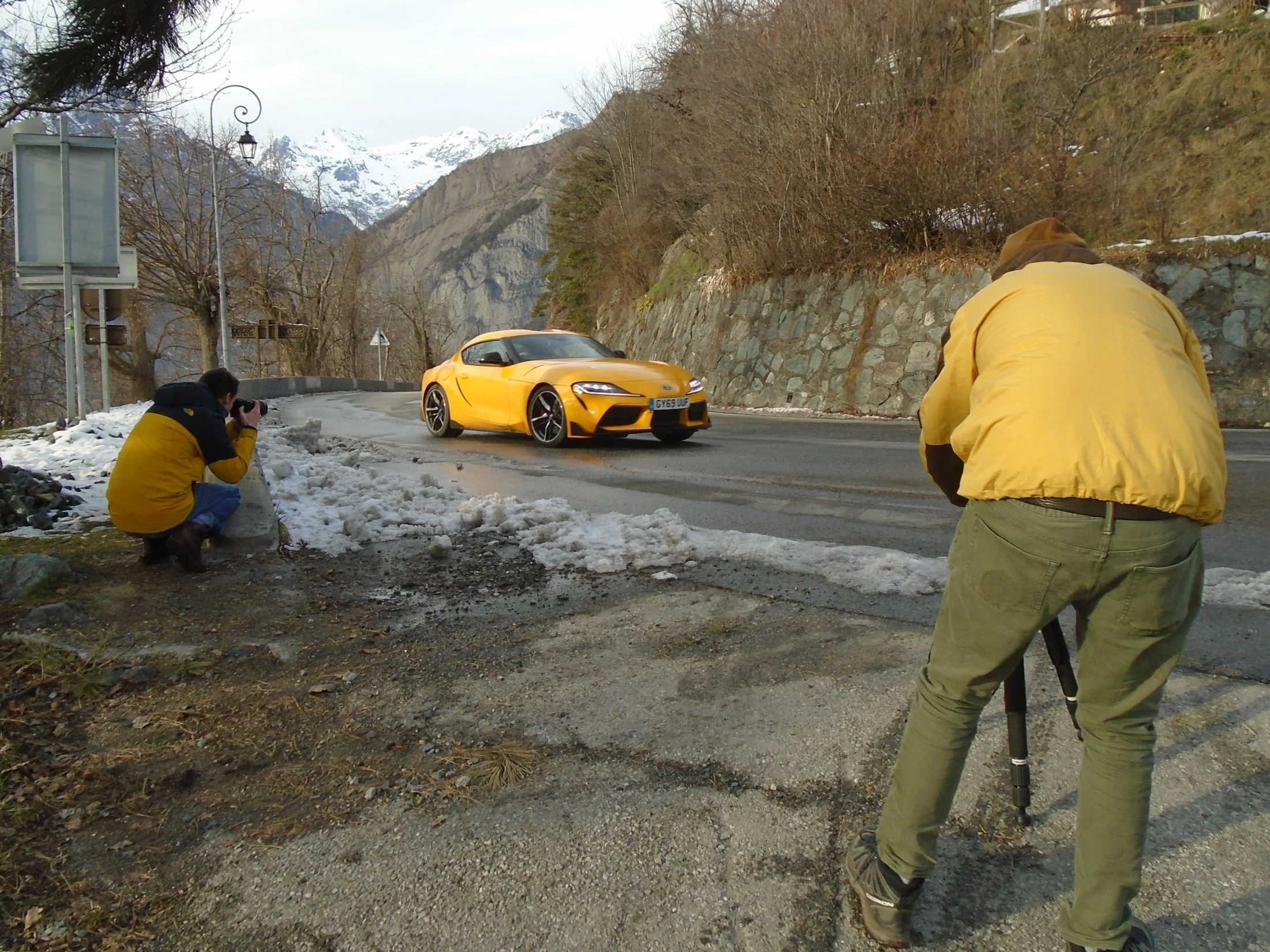 Two photographers capturing images of a yellow sports car on a mountain road with snow-capped peaks in the background.