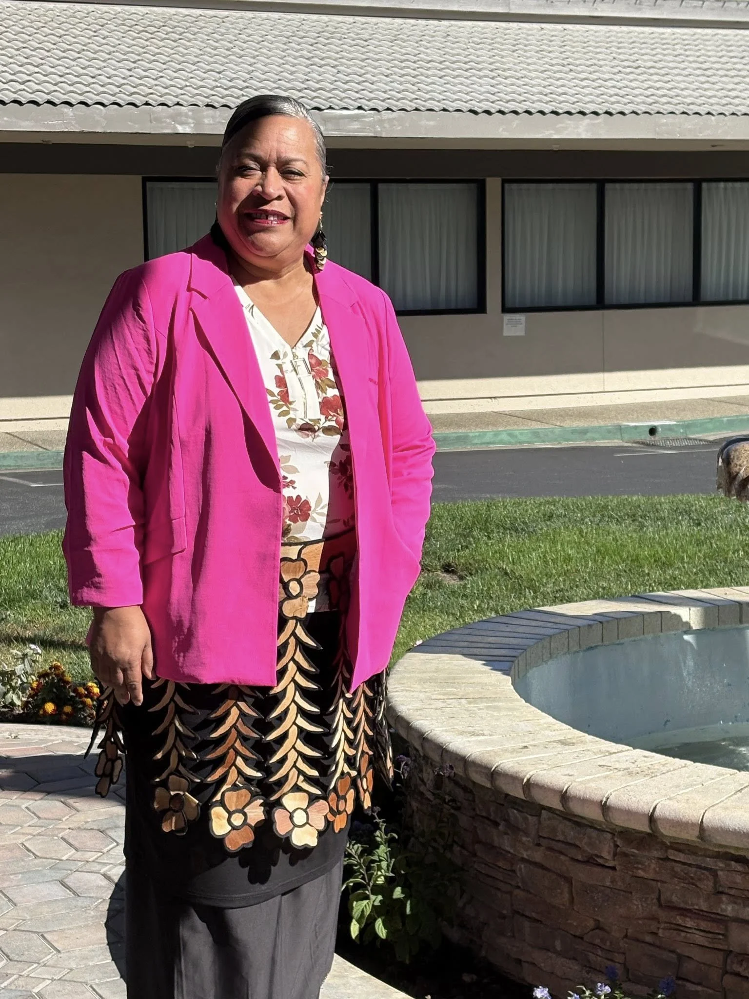 A woman standing outdoors in front of a building, wearing a bright pink blazer, a floral top, and a long skirt with a leaf and flower pattern.