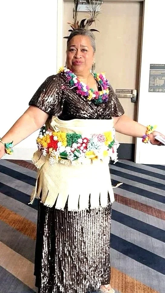 A woman is dressed in a sequined black and gold dress with a cream-colored wrap around her waist. She is wearing colorful flower leis around her neck, wrists, and waist. She stands in a hallway with striped carpet and a door behind her, making a humorous facial expression.