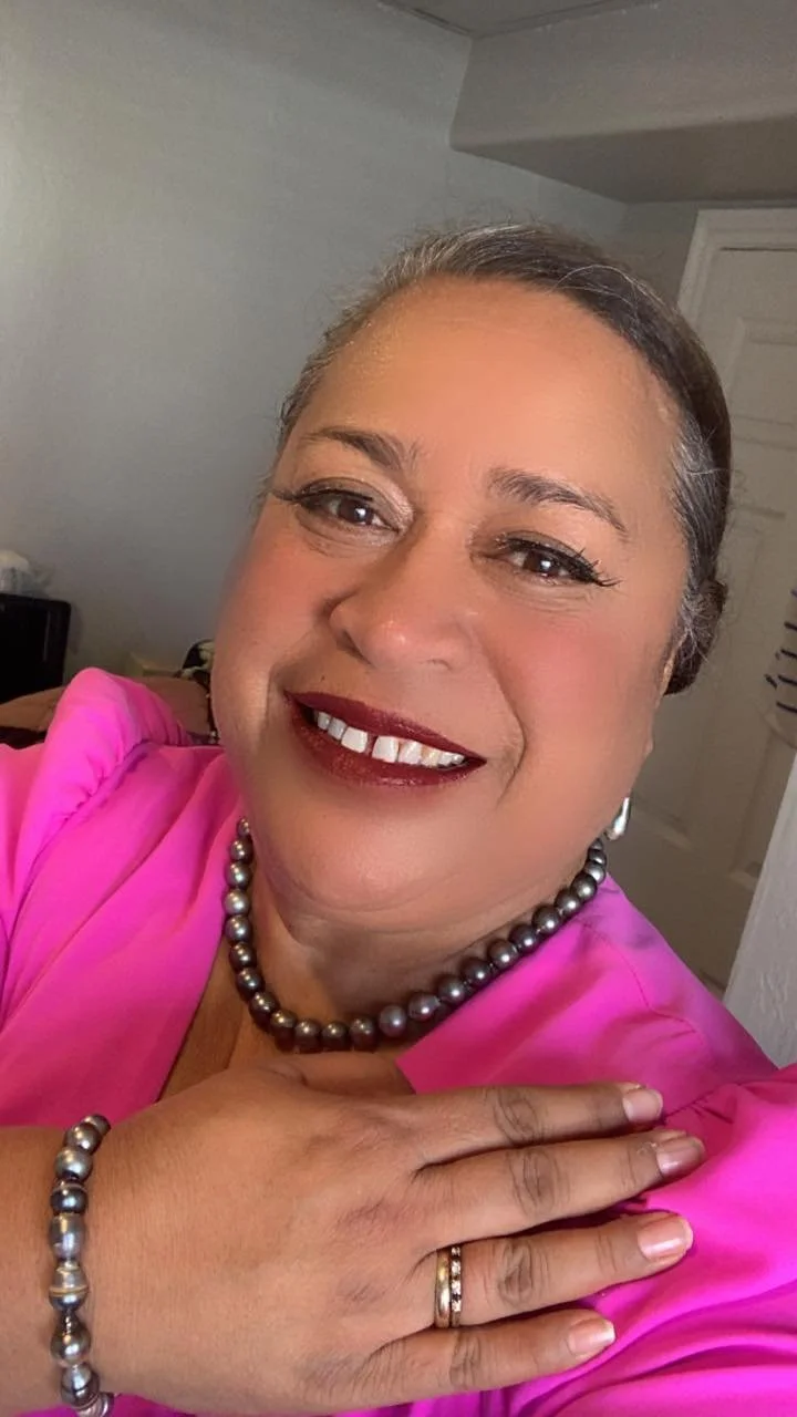A smiling middle-aged woman with gray hair and makeup, wearing a pink top, a pearl necklace, bracelet, and ring, posing with her hand on her chest and looking into the camera.
