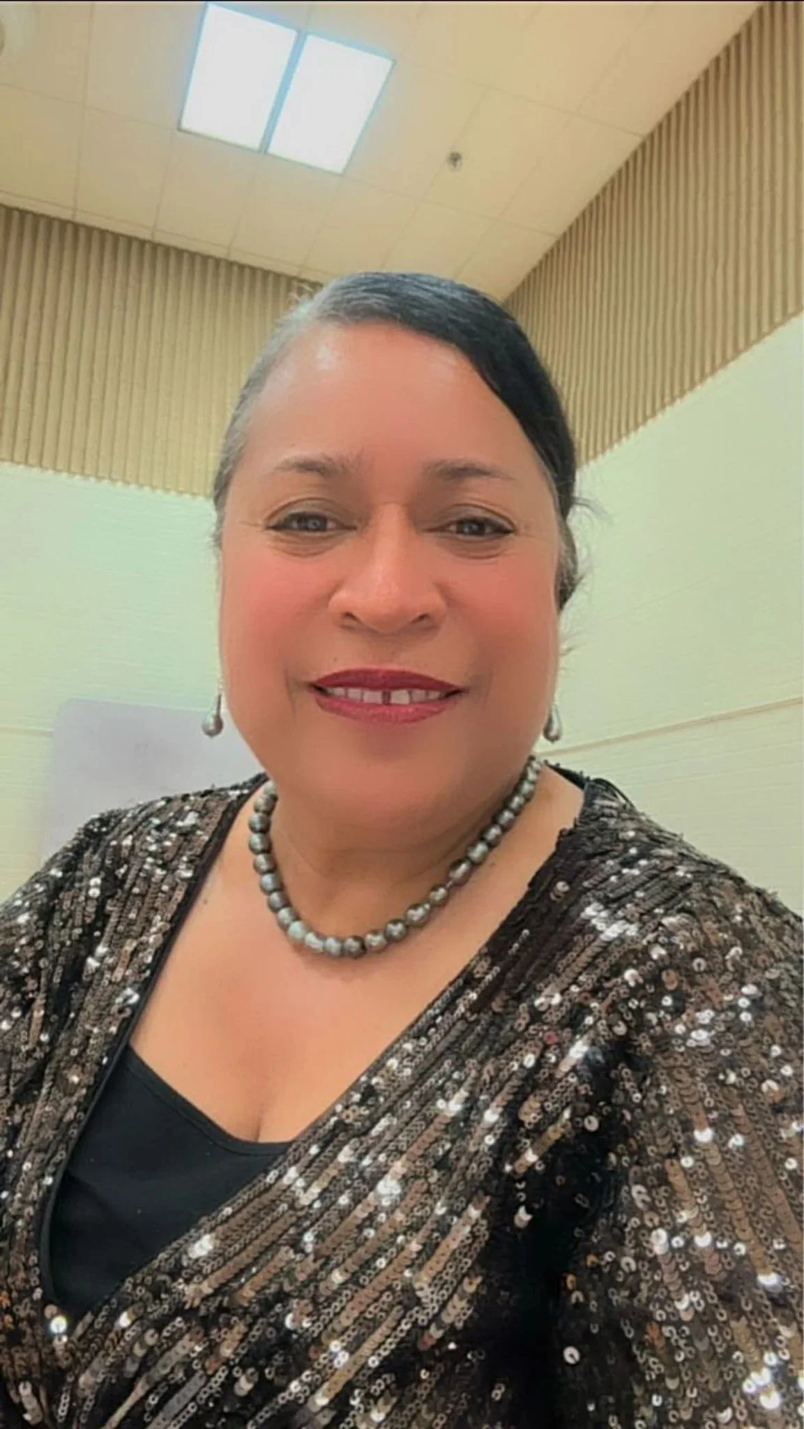 A woman with short dark hair and light makeup, smiling in a room with a beige and light green wall, wearing a sparkly black and gold top, a pearl necklace, and matching earrings.