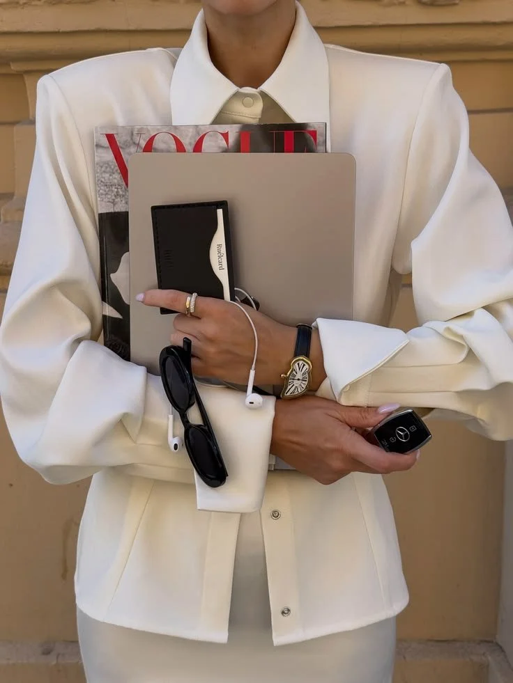 En person i en hvit blazer holder en magasin, en bok og elektroniske enheter, inkludert en smartklokke, briller og en telefon. Personen er ikke synlig over nesen.