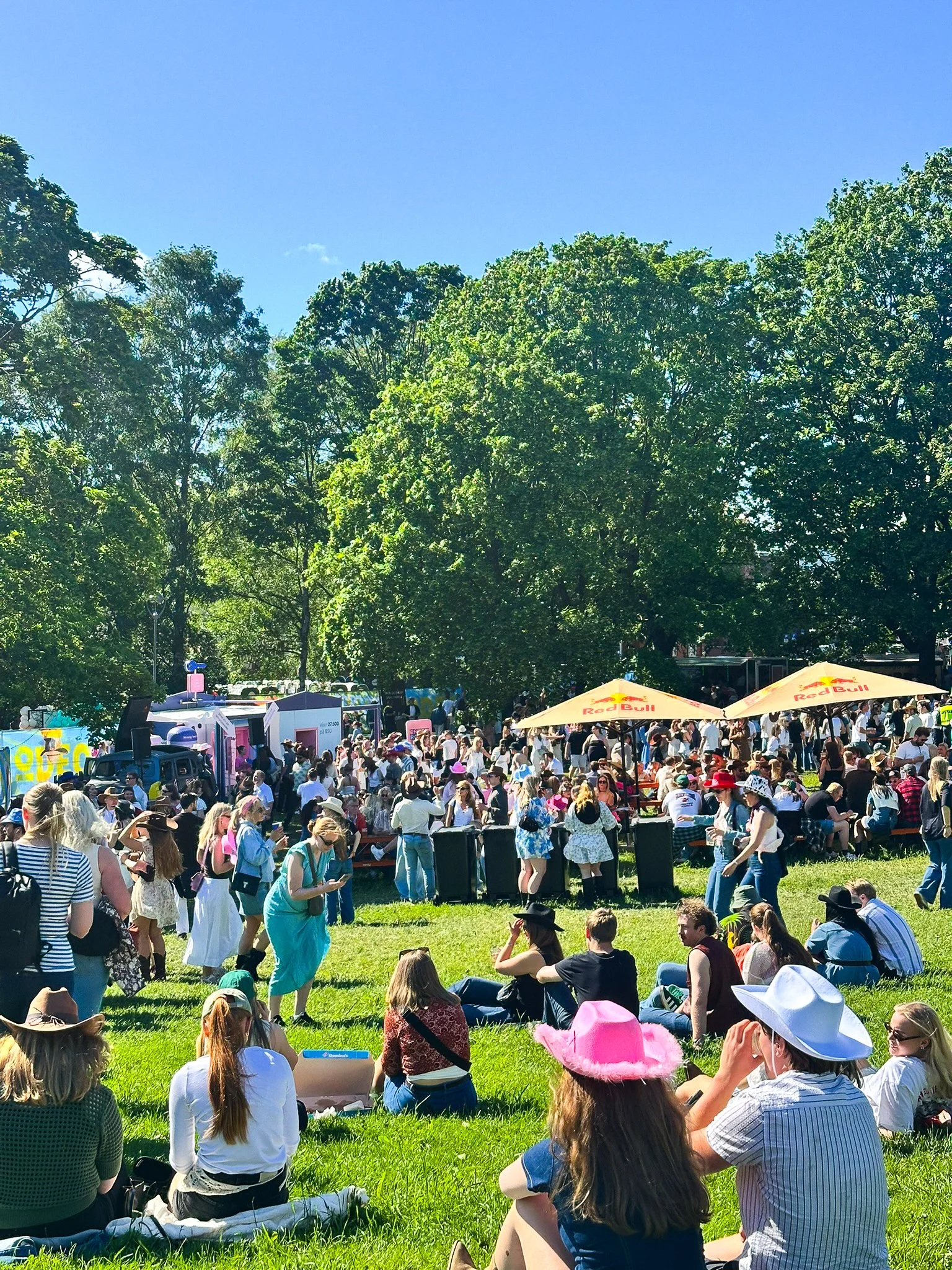 En folkemengde av mennesker samles i en park på en solskinnsdag, mange sitter på gresset og noen står eller går rundt. Det er dekorative parasoller med Red Bull-logo, og mange har på seg hatten og solbriller.