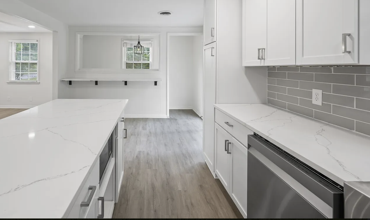 Modern white kitchen with marble countertops, gray tiled backsplash, and stainless steel dishwasher, with windows and gray wood flooring.