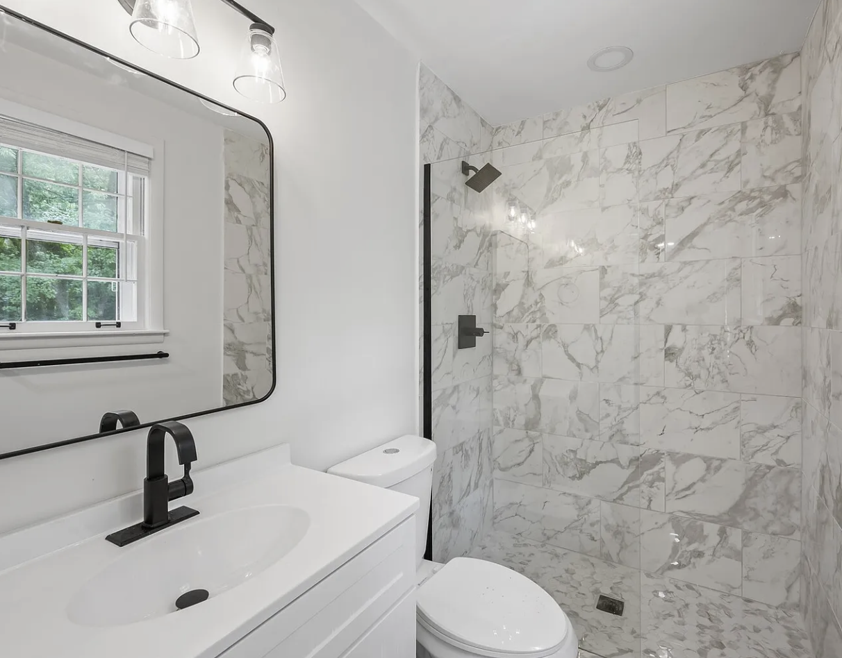 Modern bathroom with white vanity, black faucet, large mirror, toilet, and marble tiled shower area with rainfall showerhead.
