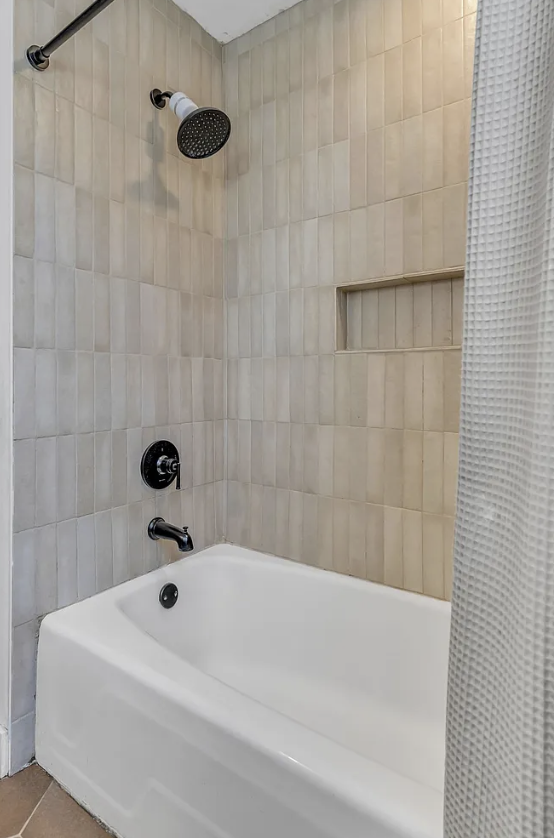 Bathroom shower area with beige tiled walls, white bathtub, black showerhead, and black control knob, with a built-in narrow shelf in the wall and a textured shower curtain partially visible.