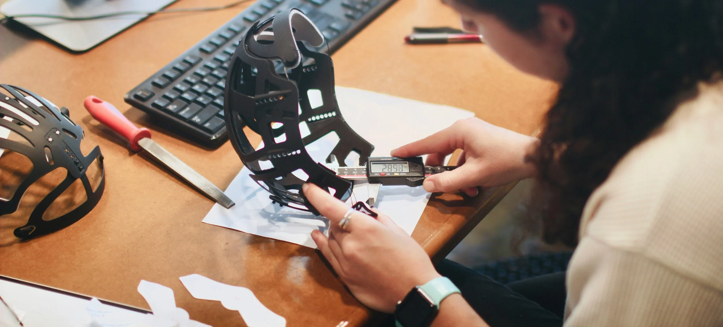 Person working on a drone frame at a desk, using a digital caliper to measure its size, surrounded by drone parts, a keyboard, and tools.