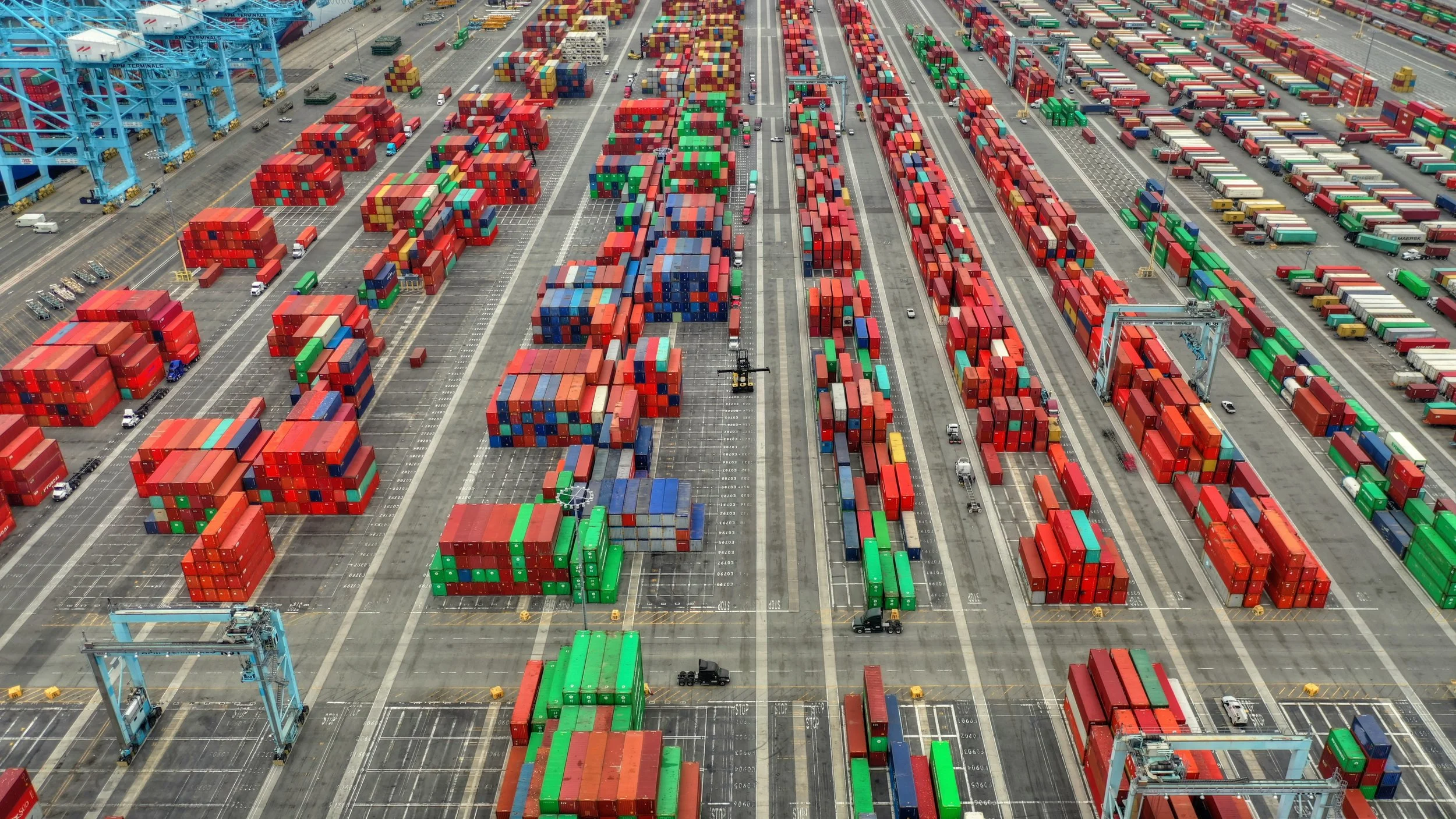 Aerial view of a shipping port with numerous stacked cargo containers in red, green, blue, and beige, along with cranes, trucks, and marked parking lanes.