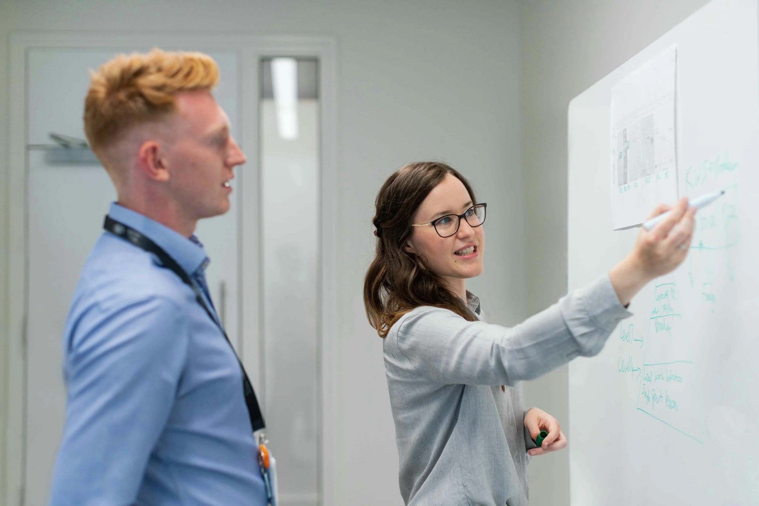 Two professionals, a man and a woman, in a modern office. The woman is writing on a whiteboard while the man looks on.