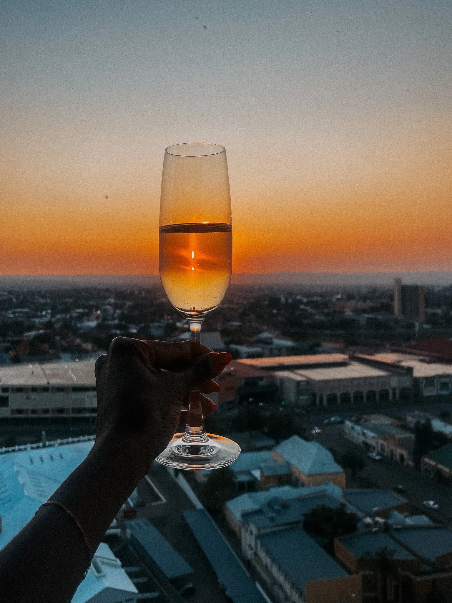 A hand holding a champagne flute filled with a golden liquid during a sunset over a cityscape.