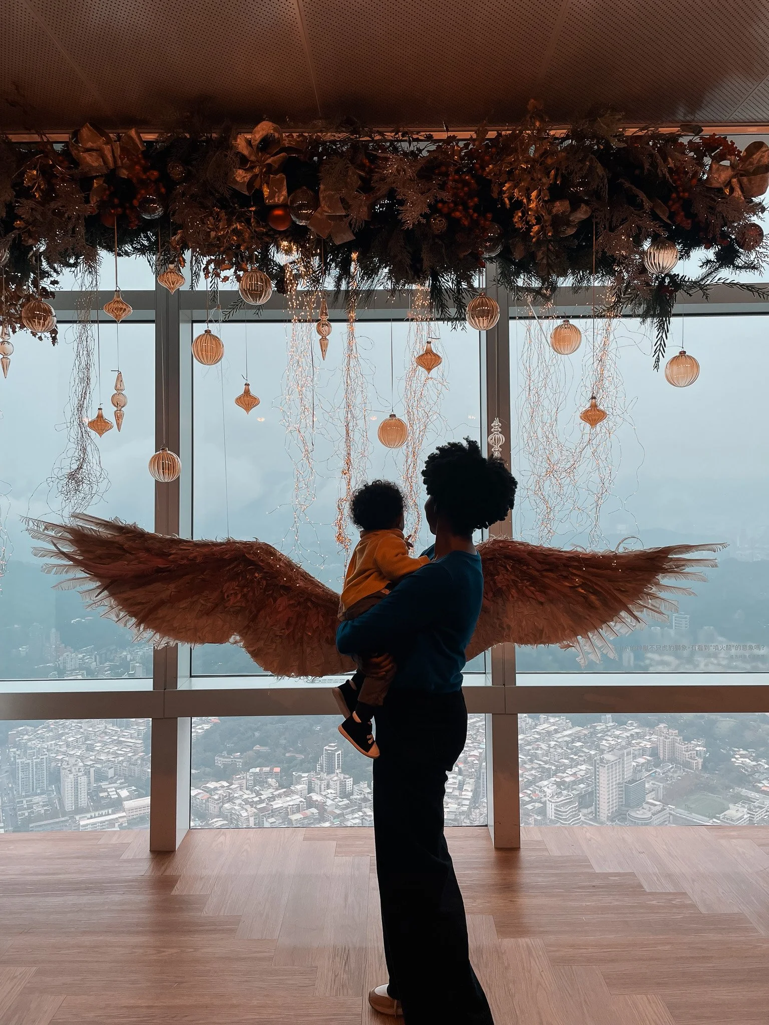 A person with angel wings holding a child in front of a large window decorated with hanging ornaments and a holiday garland.