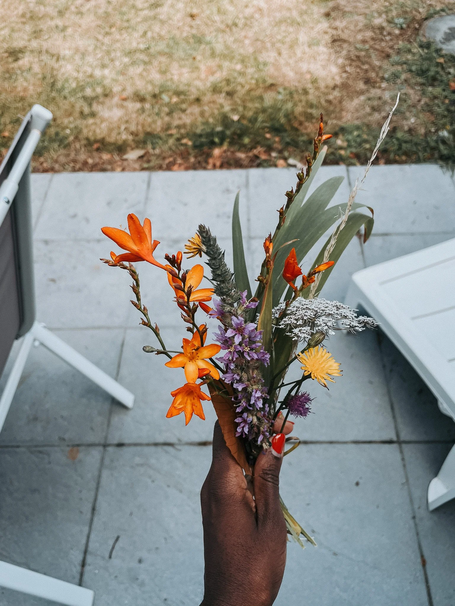 A hand holding a small bouquet of mixed flowers, including orange, purple, yellow, white, and green leaves, with an outdoor patio background.